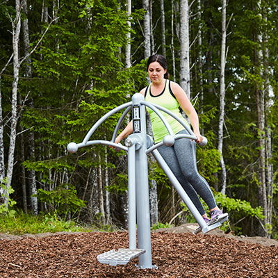 A woman is working out on an air skier at an outdoor gym surrounded with trees and nature.